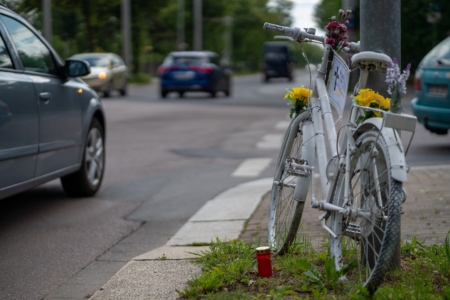 Man sieht ein weiß angemaltes Fahrrad geschmückt mit bunten Blumen an einer viel befahrenen Straße. Daneben steht eine rote Grabkerze für ein Unfallopfer.